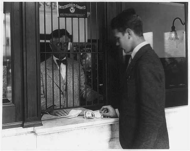 A young man in a cap slides a dollar bill across the counter at a bank teller's window, 1920s.