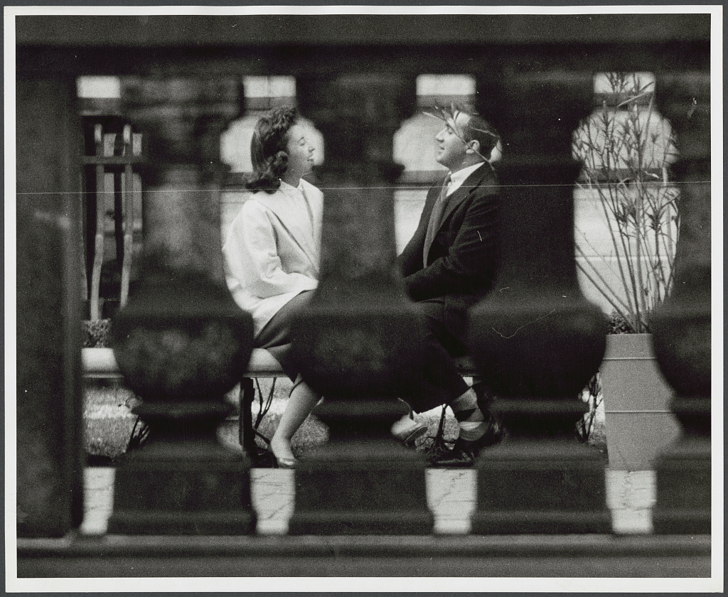 A man and a woman sit facing one another on a bench, seen through a stone railing in New York City.