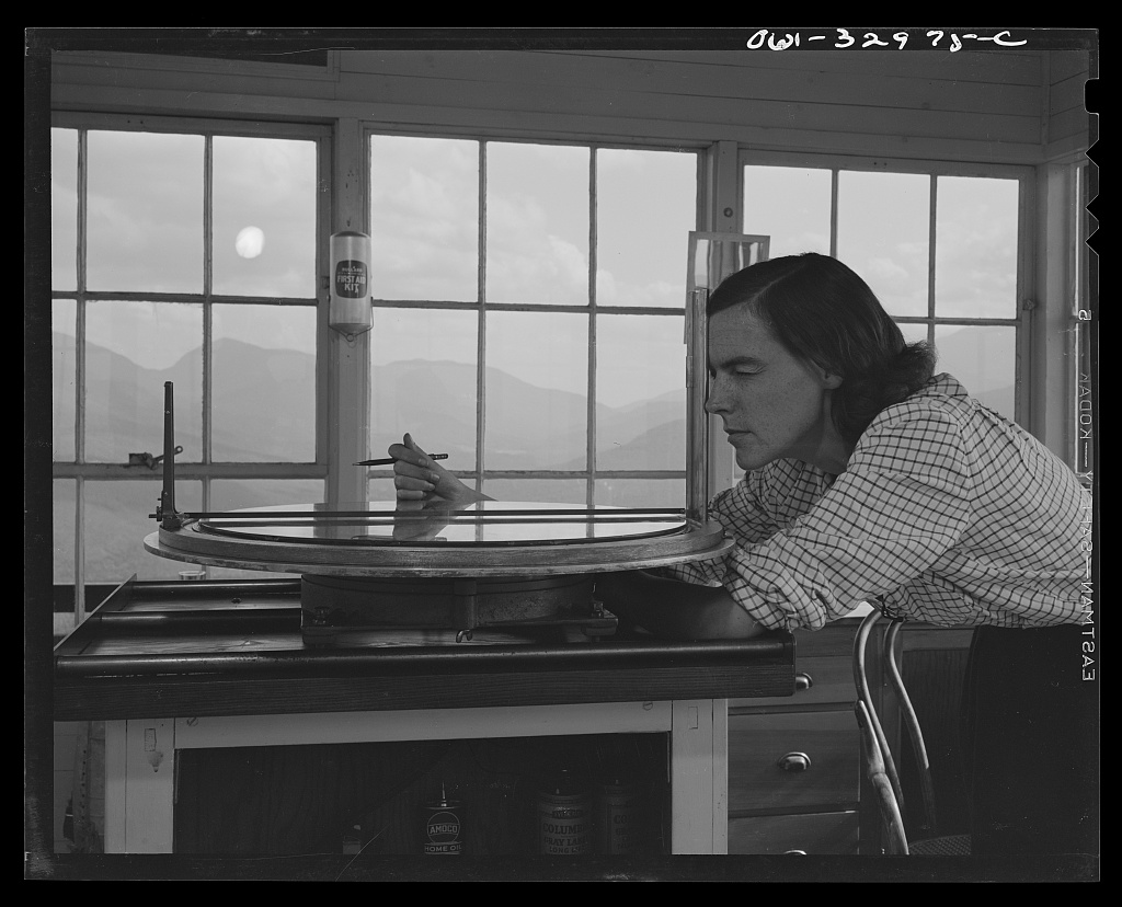 Barbara Mortensen, a fire and airplane lookout on Pine Mountain near Gorham, New Hampshire, sighting through the fire finder that locates fires on a topographical map, photographed by John Collier Jr. in June 1943